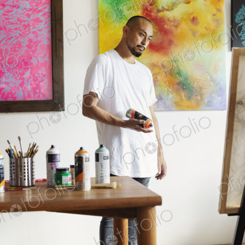 Japanese man standing in art gallery, holding can of spray paint, looking at artwork…