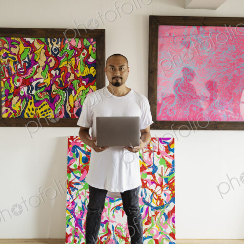 Japanese man standing in art gallery, holding laptop computer, looking at camera.