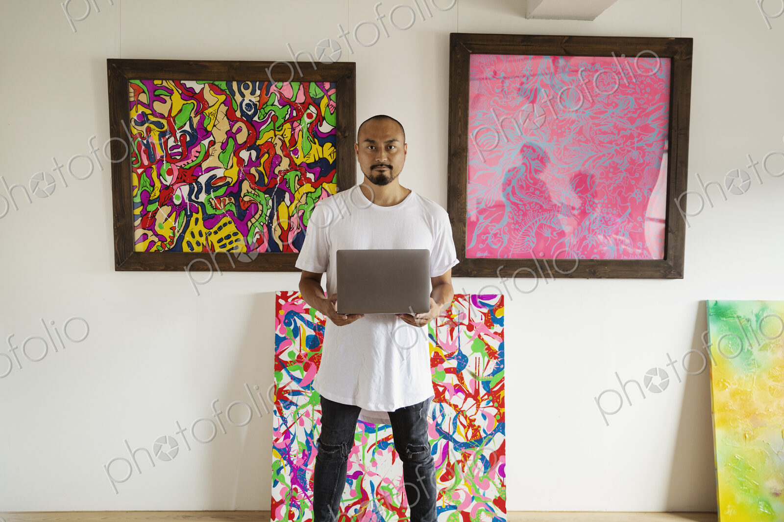 Japanese man standing in art gallery, holding laptop computer, looking at camera.