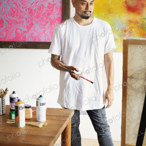 Japanese man standing in art gallery, holding paintbrush, looking at artwork on easel.