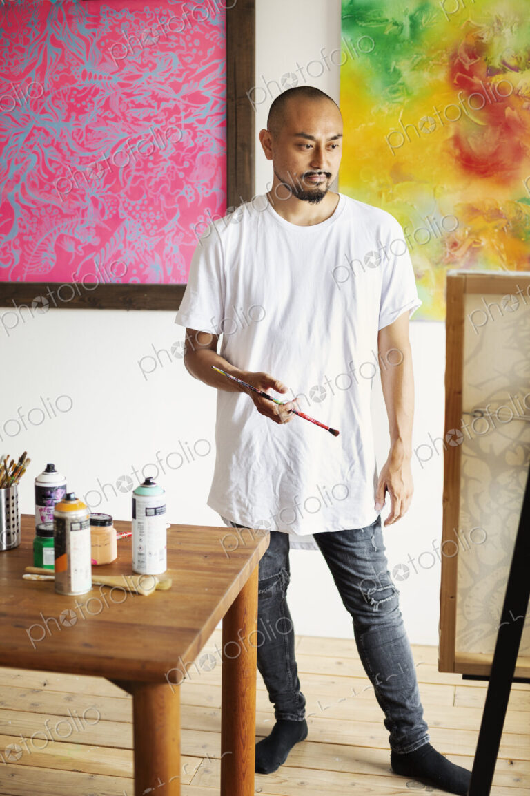 Japanese man standing in art gallery, holding paintbrush, looking at artwork on easel.