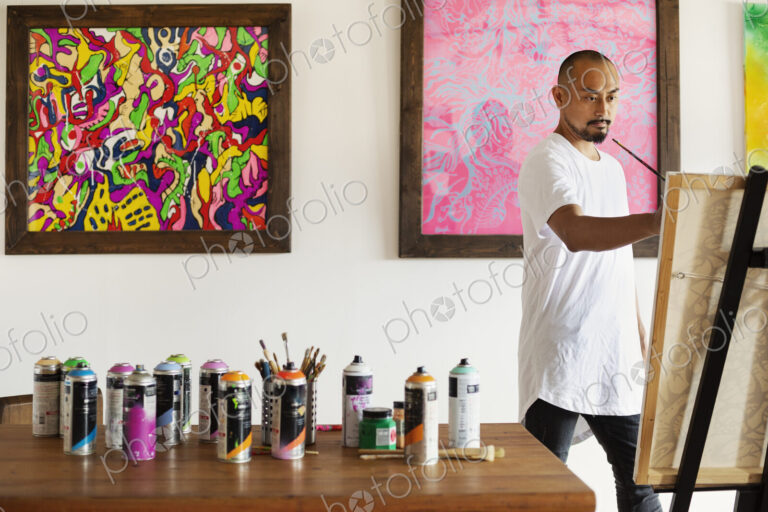 Japanese man standing in art gallery, holding paintbrush, working on artwork on easel.