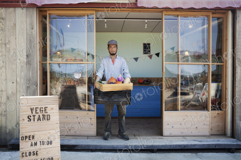 Japanese man standing outside a farm shop, holding crate with fresh vegetables,…