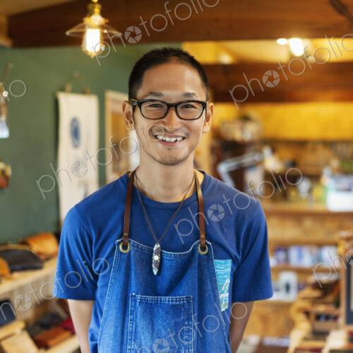 Japanese man wearing blue apron and glasses standing in a leather shop, smiling at camera.