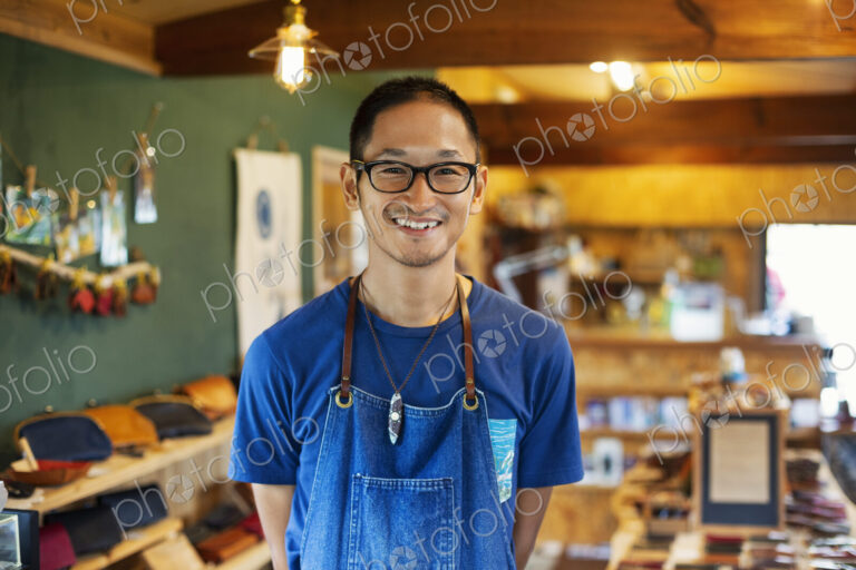 Japanese man wearing blue apron and glasses standing in a leather shop, smiling at camera.