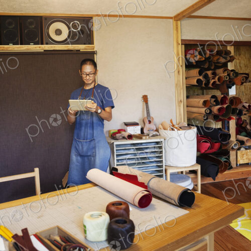 Japanese man wearing blue apron and glasses standing in a leather shop, using digital… Japanese man wearing blue apron and glasses standing in a leather shop, using digital…