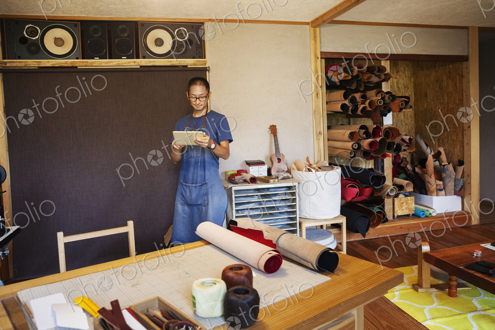 Japanese man wearing blue apron and glasses standing in a leather shop, using digital…