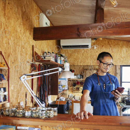 Japanese man wearing blue apron and glasses standing in a leather shop, using his… Japanese man wearing blue apron and glasses standing in a leather shop, using his…