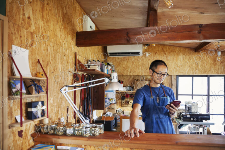 Japanese man wearing blue apron and glasses standing in a leather shop, using his…