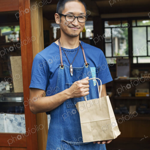 Japanese man wearing blue apron and glasses standing outside a leather shop, holding… Japanese man wearing blue apron and glasses standing outside a leather shop, holding…