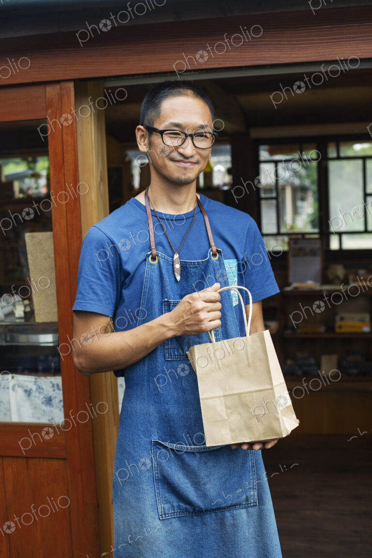 Japanese man wearing blue apron and glasses standing outside a leather shop, holding…