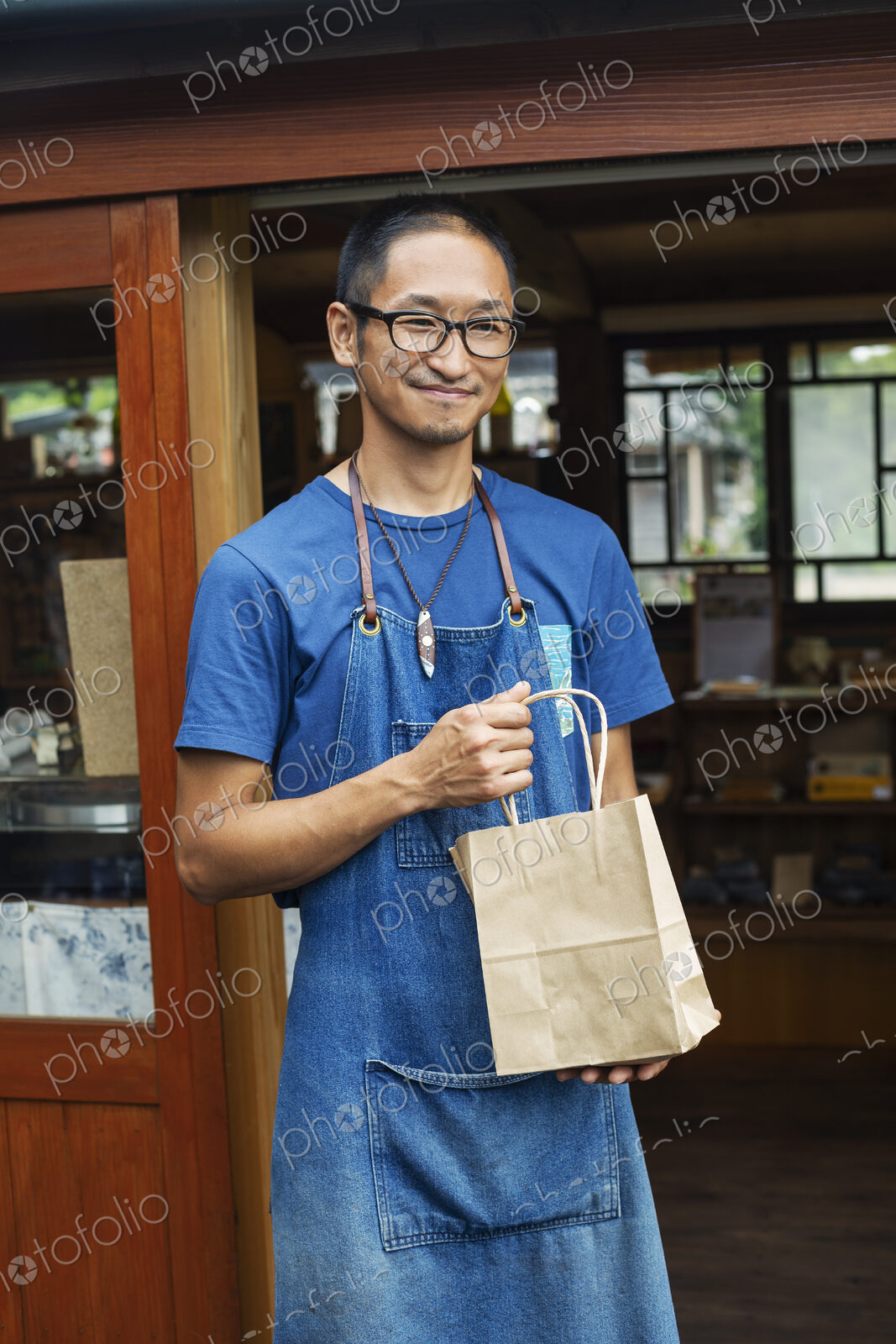 Japanese man wearing blue apron and glasses standing outside a leather shop, holding…
