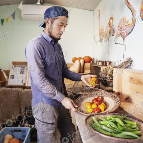 Japanese man wearing cap standing in farm shop, holding bowl with fresh vegetables.