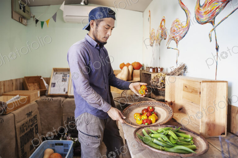Japanese man wearing cap standing in farm shop, holding bowl with fresh vegetables.