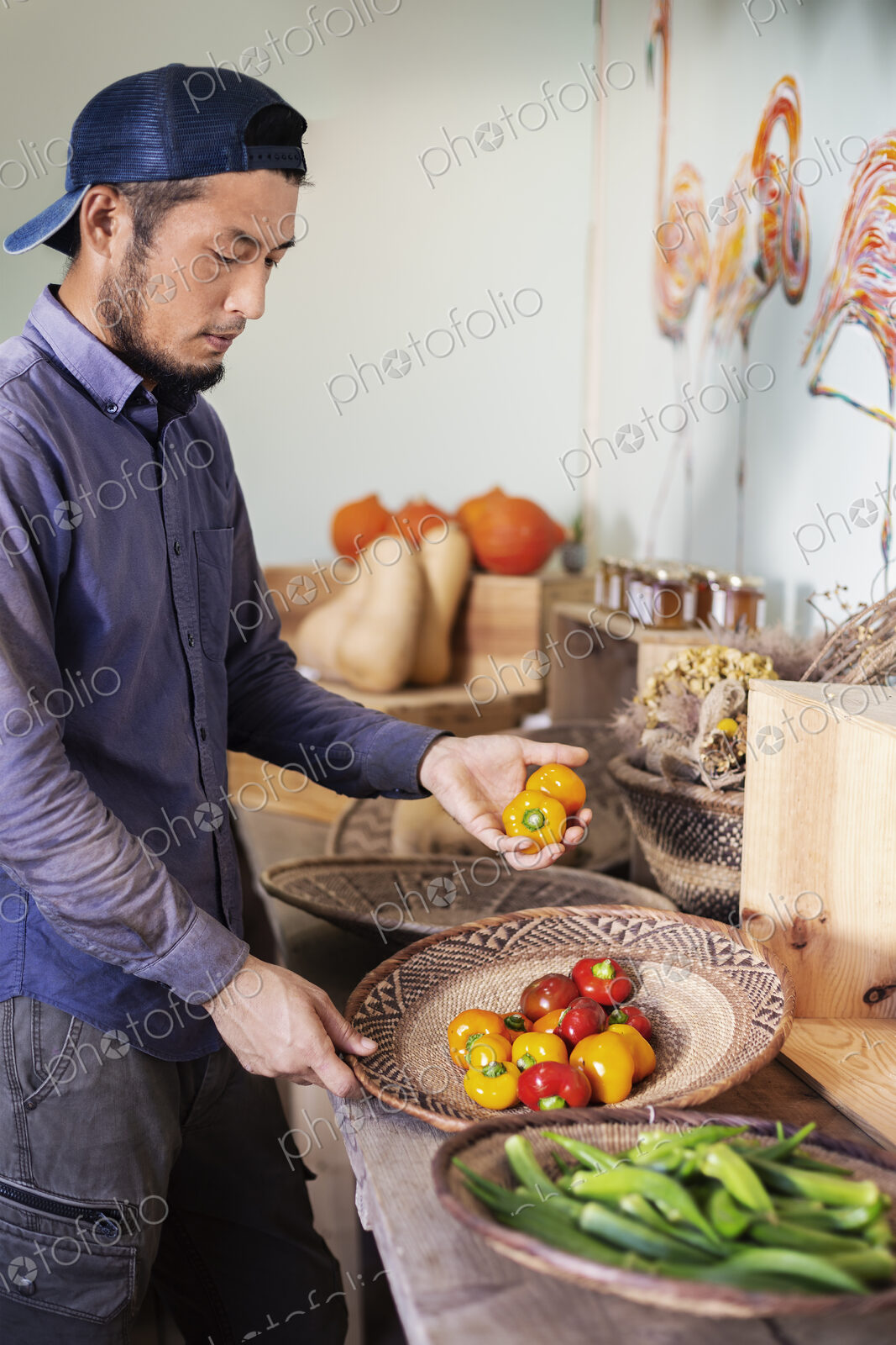Japanese man wearing cap standing in farm shop, holding bowl with fresh vegetables.