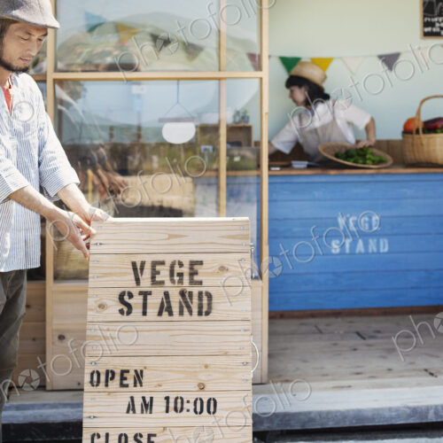 Japanese man wearing cap standing outside a farm shop, putting up wooden shop sign.