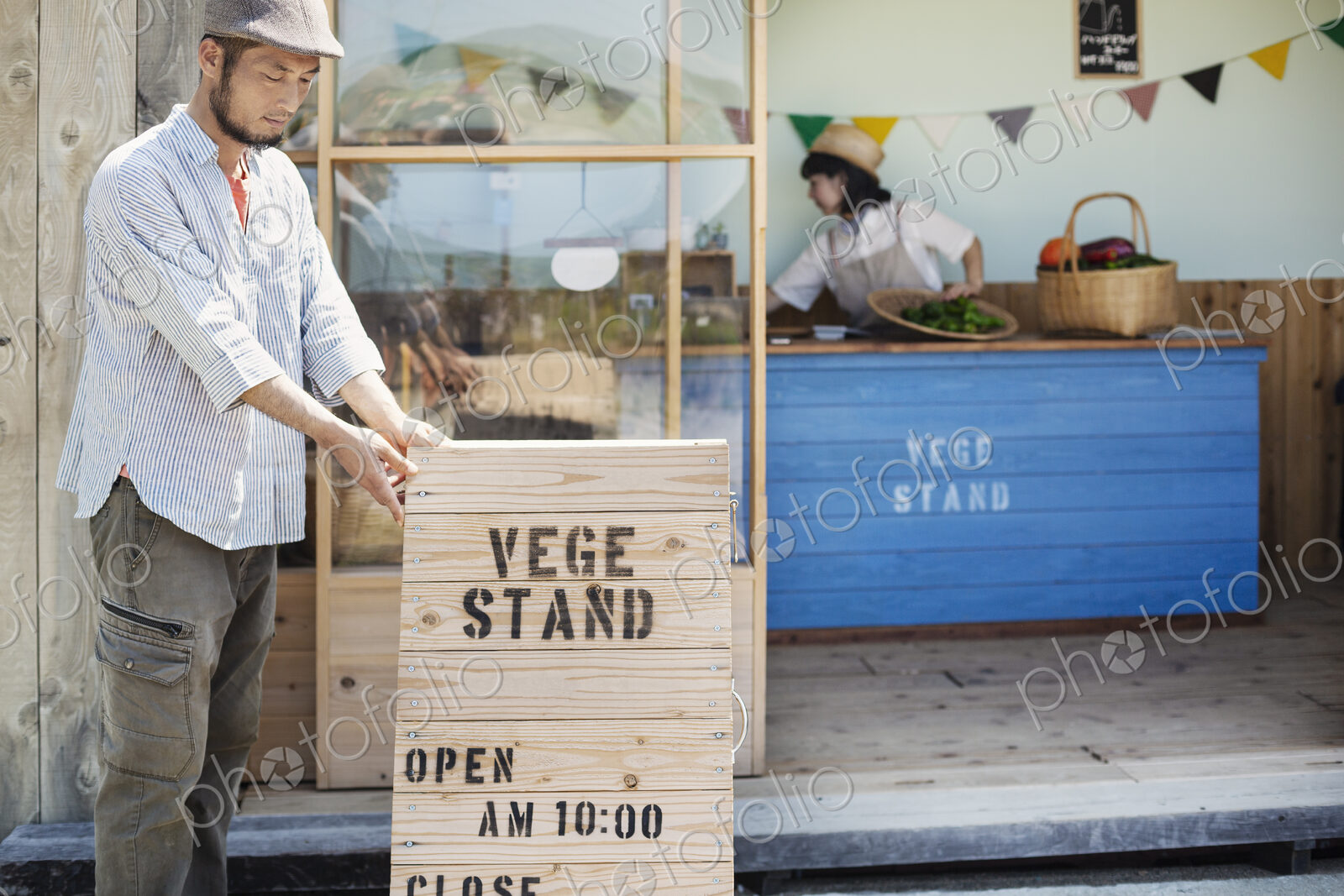 Japanese man wearing cap standing outside a farm shop, putting up wooden shop sign.