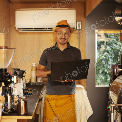 Japanese man wearing hat and glasses standing in an Eco Cafe, holding laptop computer,… Japanese man wearing hat and glasses standing in an Eco Cafe, holding laptop computer,…