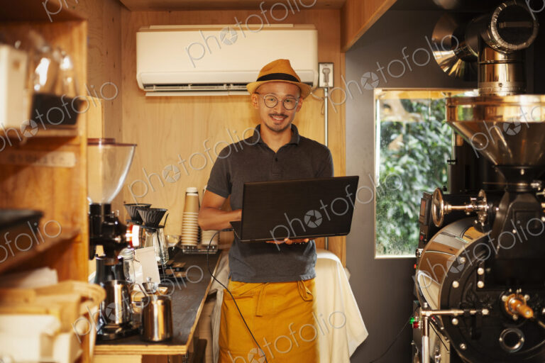 Japanese man wearing hat and glasses standing in an Eco Cafe, holding laptop computer,…