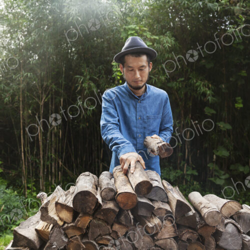 Japanese man wearing hat standing outdoors, stacking logs of firewood. Japanese man wearing hat standing outdoors, stacking logs of firewood.