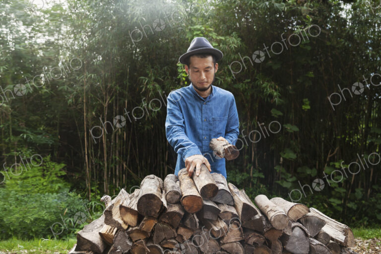 Japanese man wearing hat standing outdoors, stacking logs of firewood.