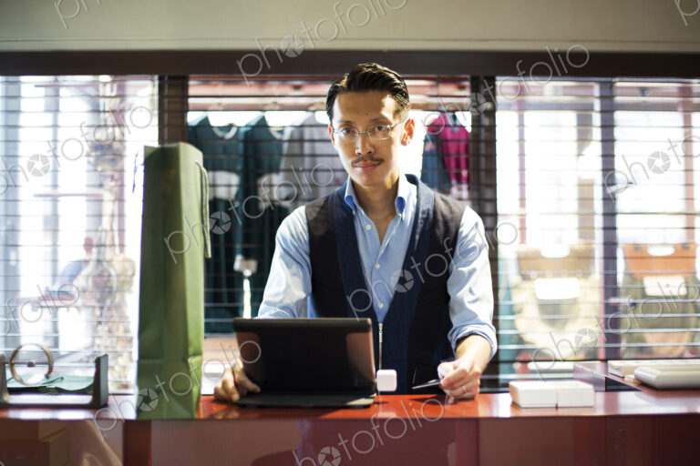 Japanese salesman with moustache wearing glasses standing at counter in clothing…