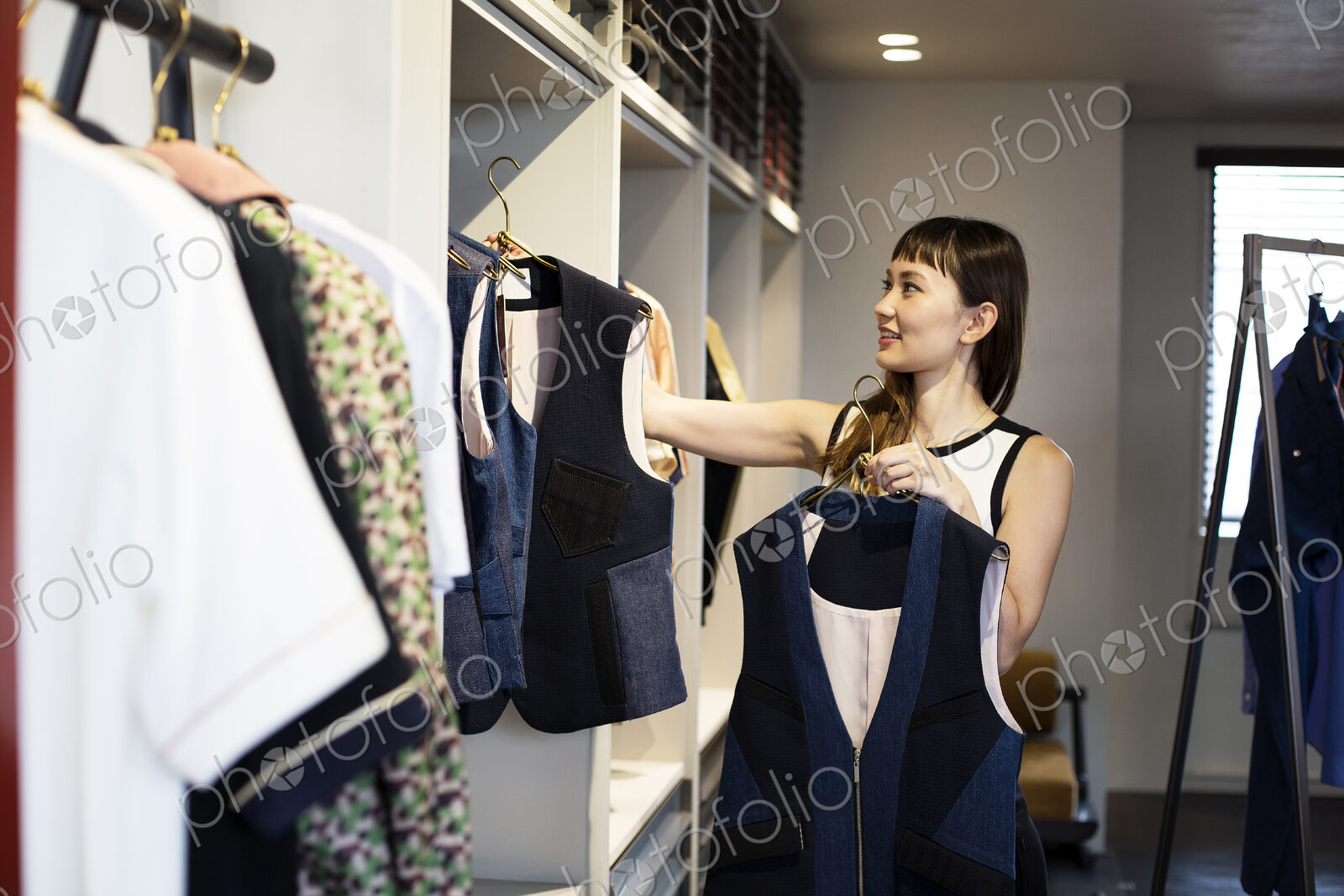 Japanese saleswoman standing in clothing store, hanging blue waistcoats on rail.
