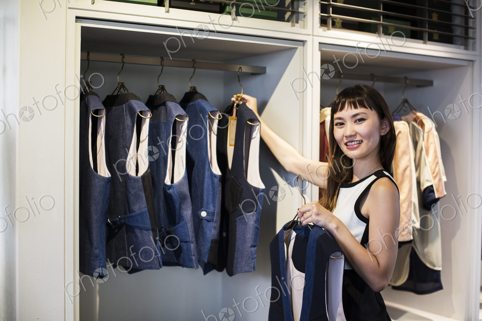 Japanese saleswoman standing in clothing store, hanging blue waistcoats on rail,…