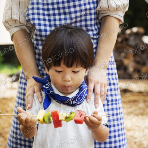 Japanese woman and boy standing outdoors, holding skewer with fresh kiwi and melon.