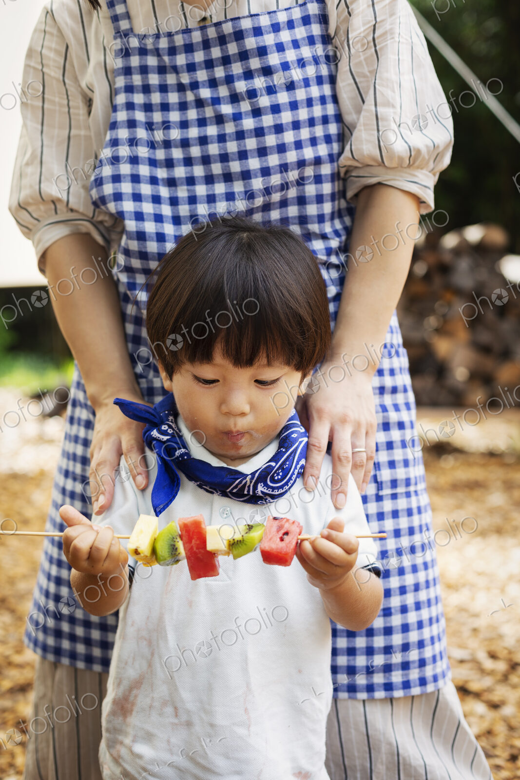 Japanese woman and boy standing outdoors, holding skewer with fresh kiwi and melon.