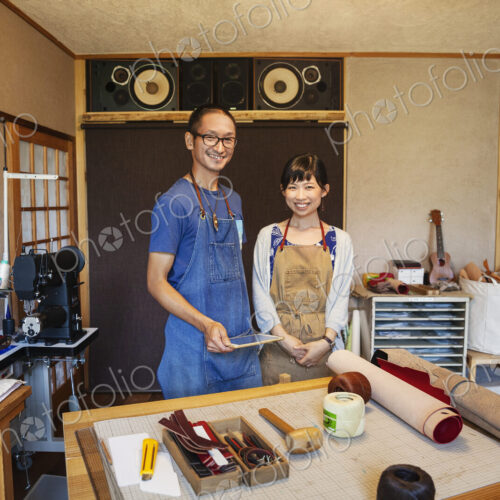 Japanese woman and man wearing blue apron and glasses standing in a leather shop,…