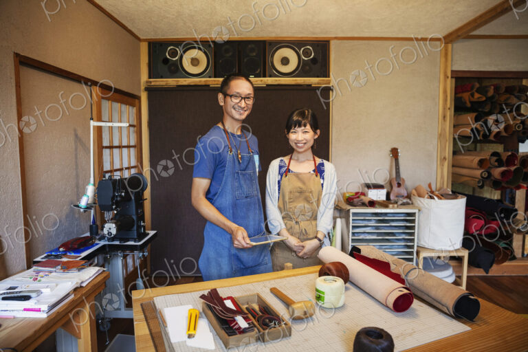 Japanese woman and man wearing blue apron and glasses standing in a leather shop,…