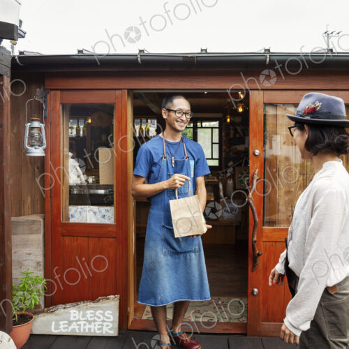 Japanese woman and man wearing blue apron and glasses standing outside a leather shop,… Japanese woman and man wearing blue apron and glasses standing outside a leather shop,…