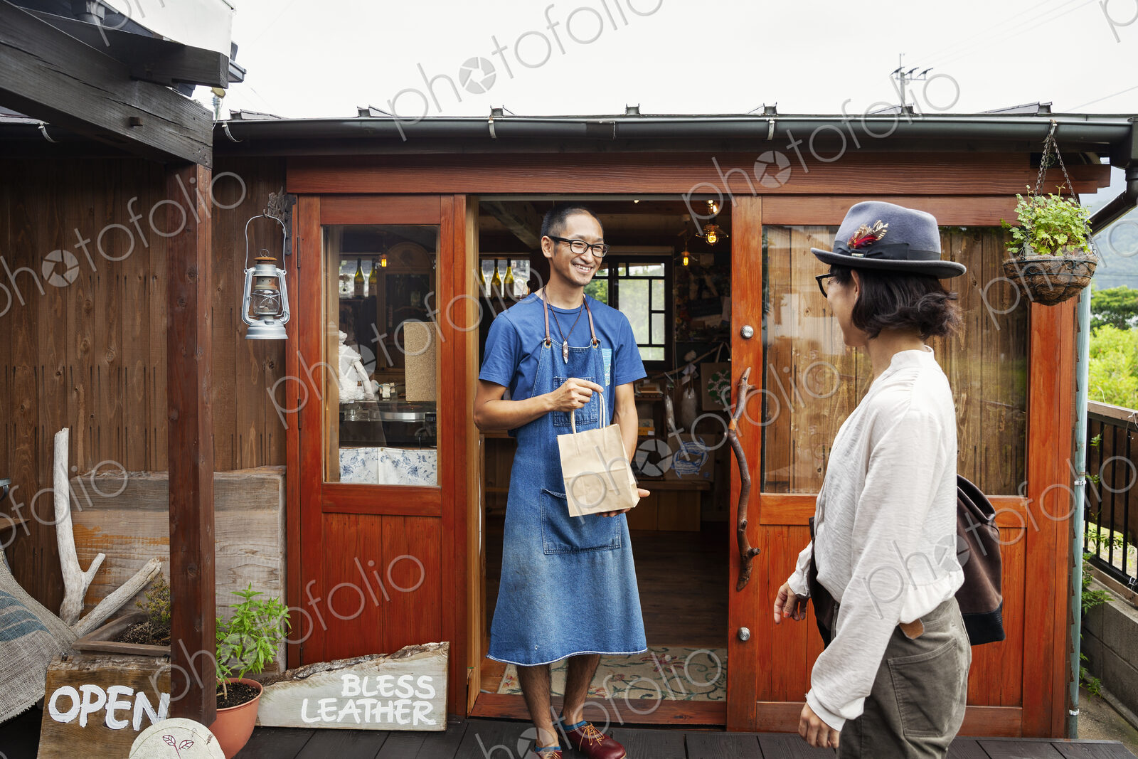 Japanese woman and man wearing blue apron and glasses standing outside a leather shop,…