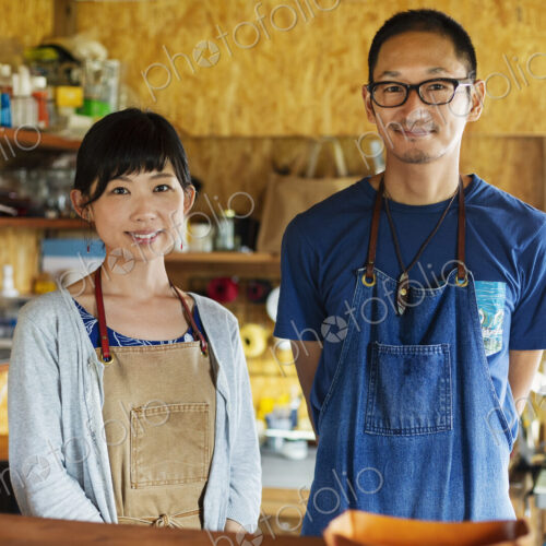Japanese woman and man wearing blue apron standing in a leather shop, smiling at camera.