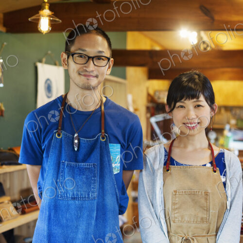 Japanese woman and man wearing blue apron standing in a leather shop, smiling at camera.