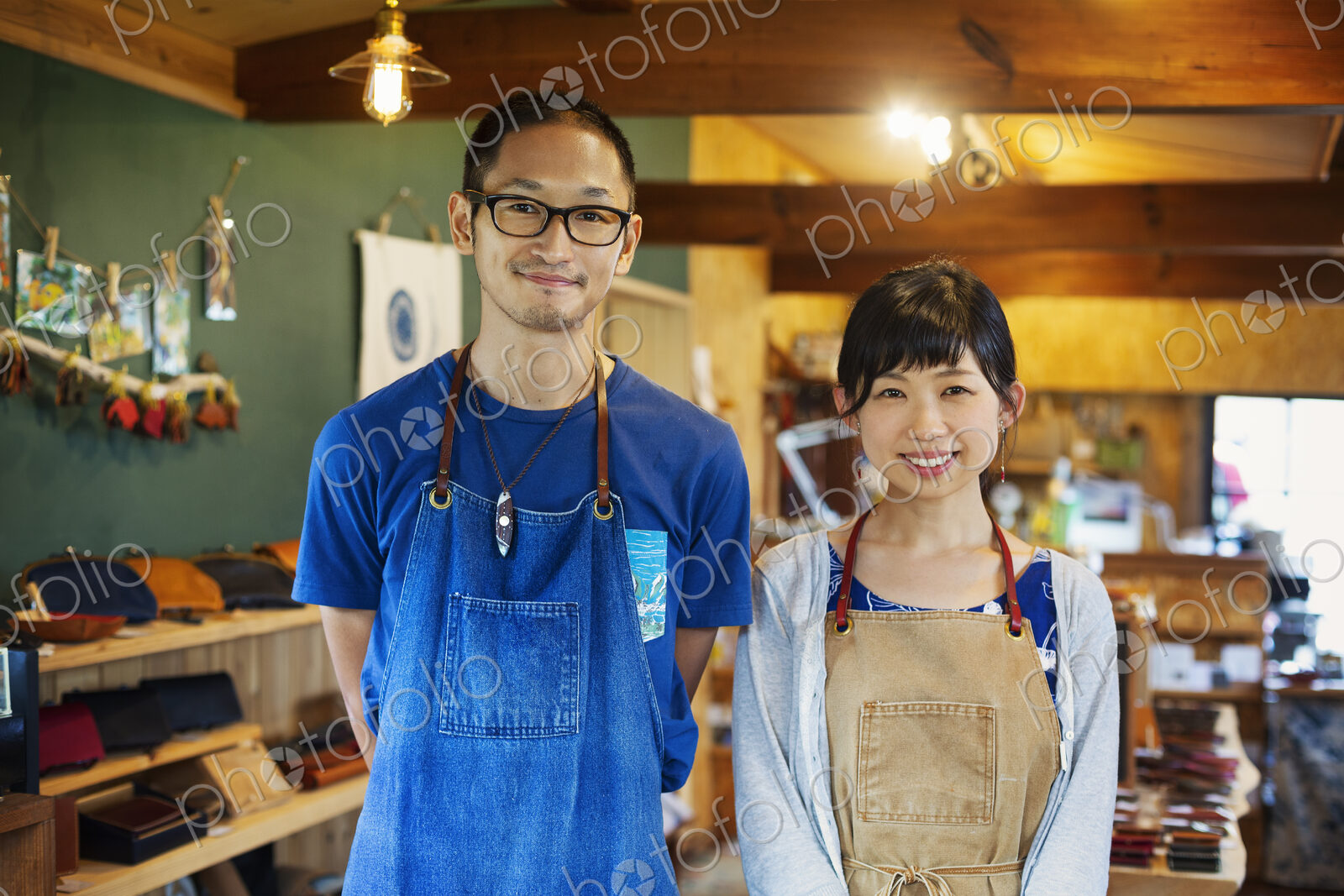 Japanese woman and man wearing blue apron standing in a leather shop, smiling at camera.