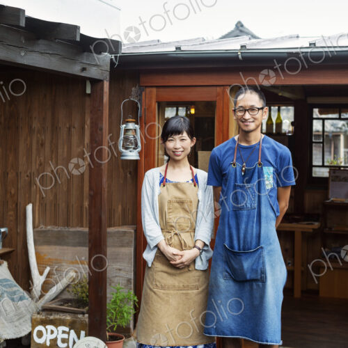 Japanese woman and man wearing blue apron standing outside a leather shop, smiling at…