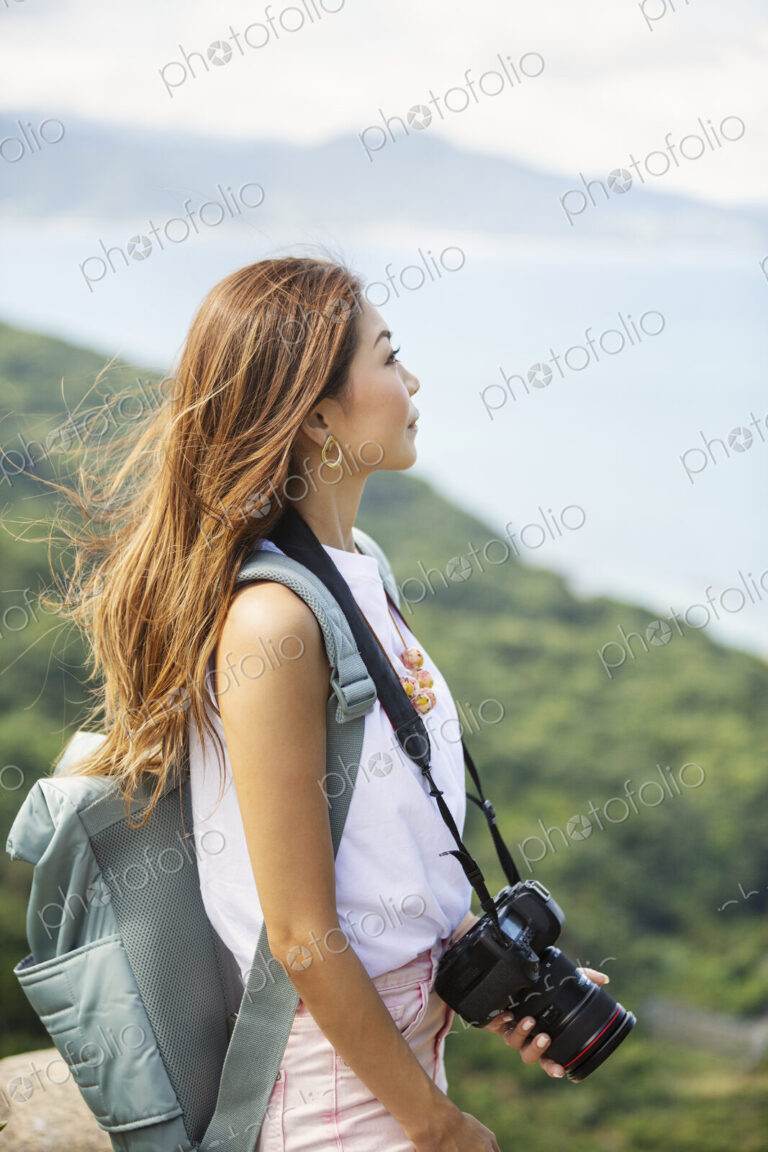 Japanese woman carrying backpack and holding camera standing on a cliff, ocean in the…