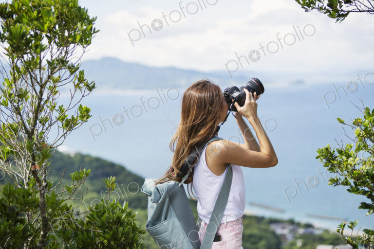 Japanese woman carrying backpack taking photograph on a cliff, ocean in the background.