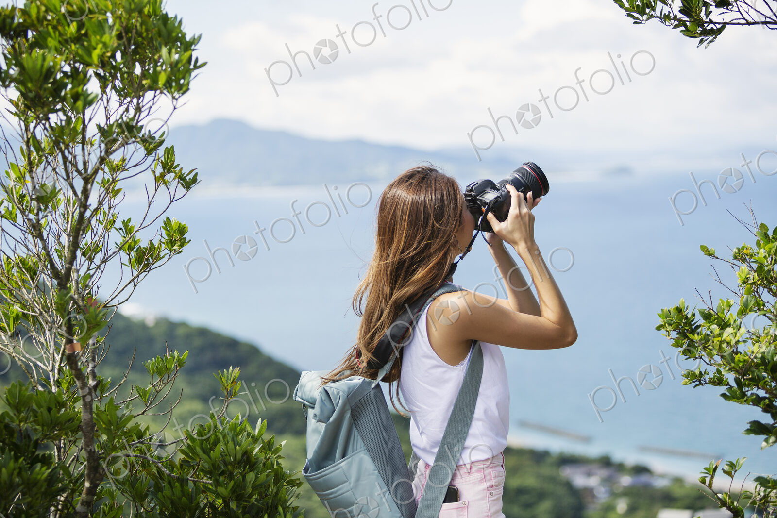 Japanese woman carrying backpack taking photograph on a cliff, ocean in the background.
