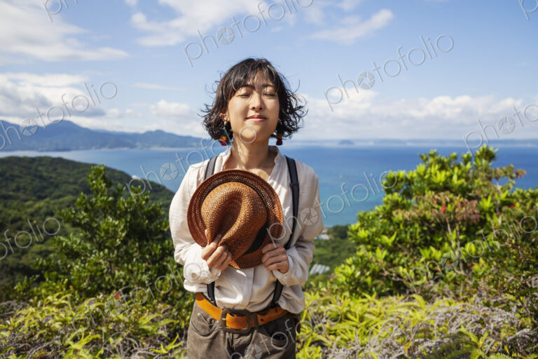 Japanese woman holding hat standing on a cliff, ocean in the background.