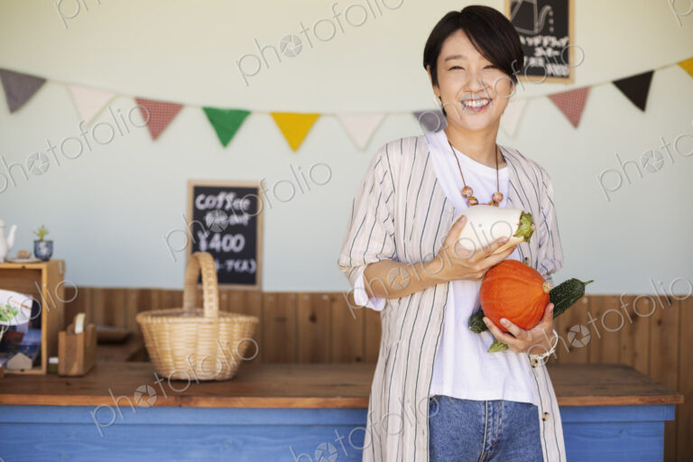 Japanese woman standing in a farm shop, holding vegetables, smiling at camera.
