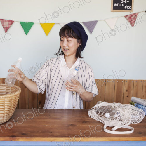 Japanese woman standing in a farm shop, sorting clear plastic bottles into basket.