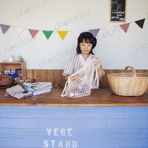 Japanese woman standing in a farm shop, sorting clear plastic bottles into net bag and…