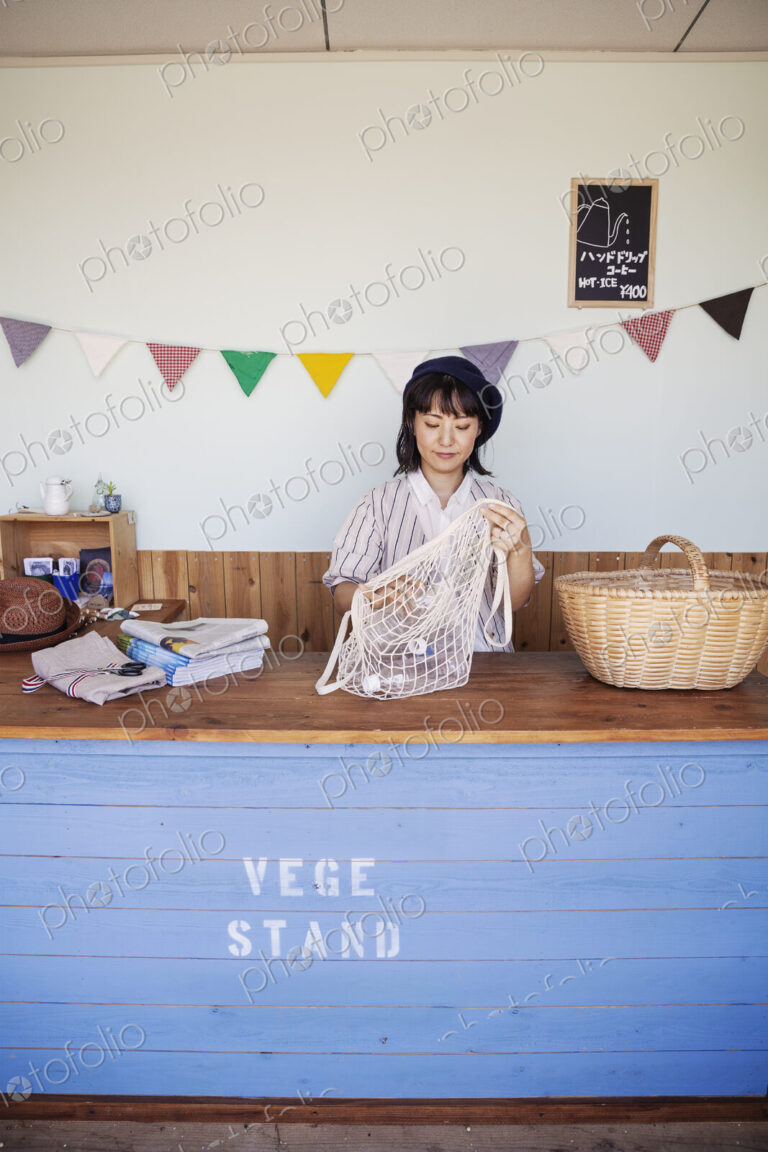 Japanese woman standing in a farm shop, sorting clear plastic bottles into net bag and…