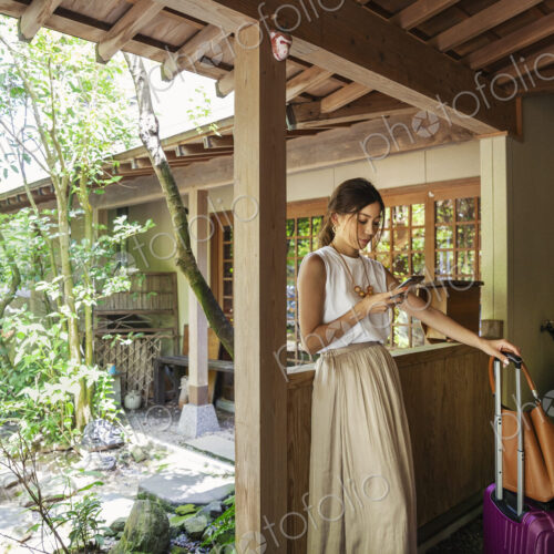 Japanese woman standing on a porch, using mobile phone, holding suitcase and handbag.