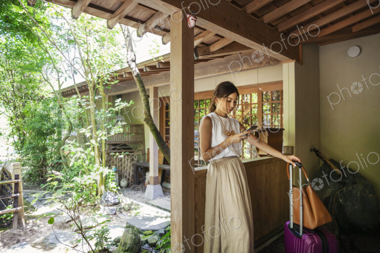 Japanese woman standing on a porch, using mobile phone, holding suitcase and handbag.
