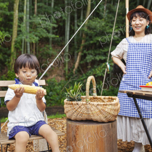 Japanese woman standing outdoors, wearing hat and apron and young boy sitting on…