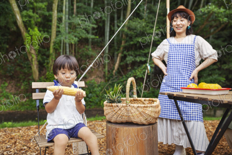 Japanese woman standing outdoors, wearing hat and apron and young boy sitting on…